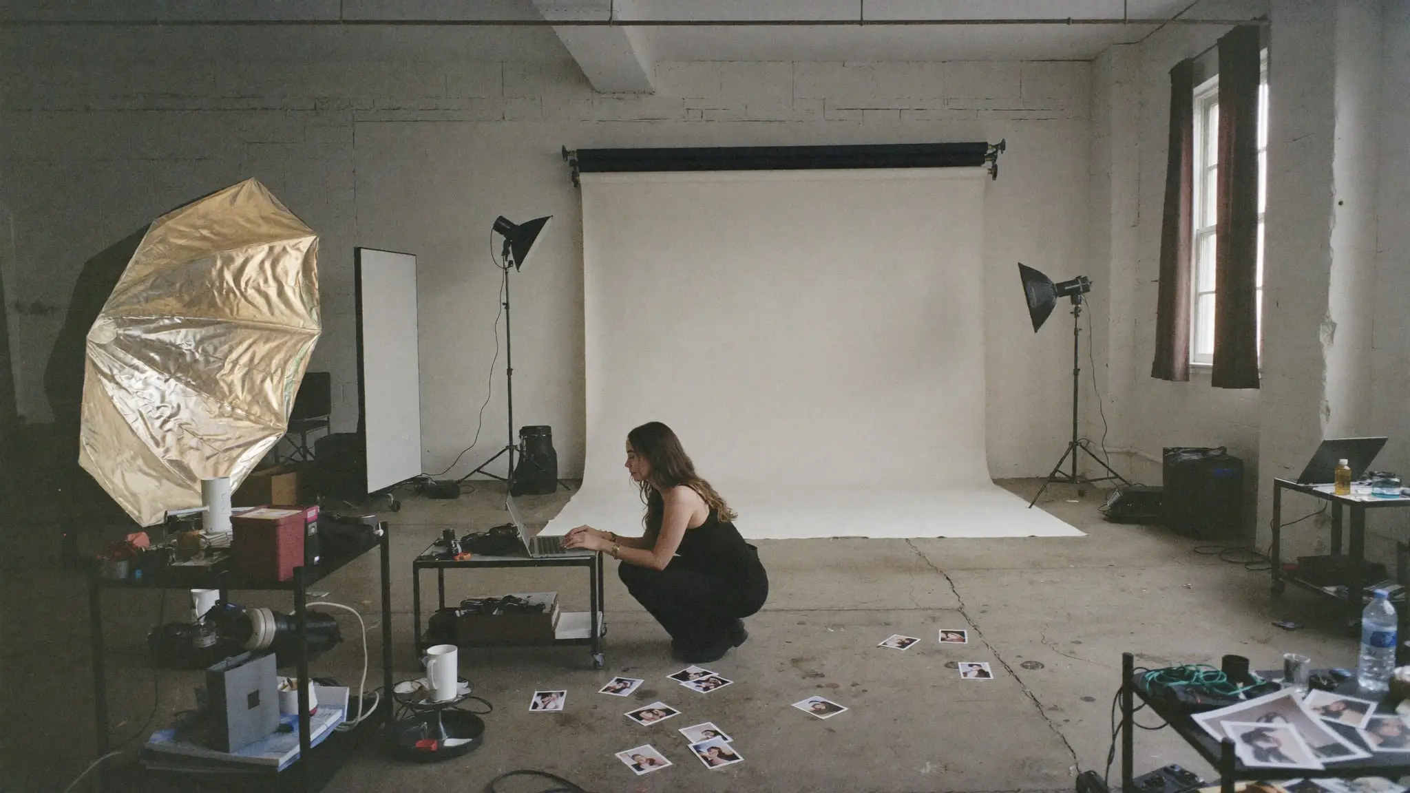 Paige Pope crouched at a laptop in a working photo studio, with lights, a paper backdrop and prints laid out on the floor