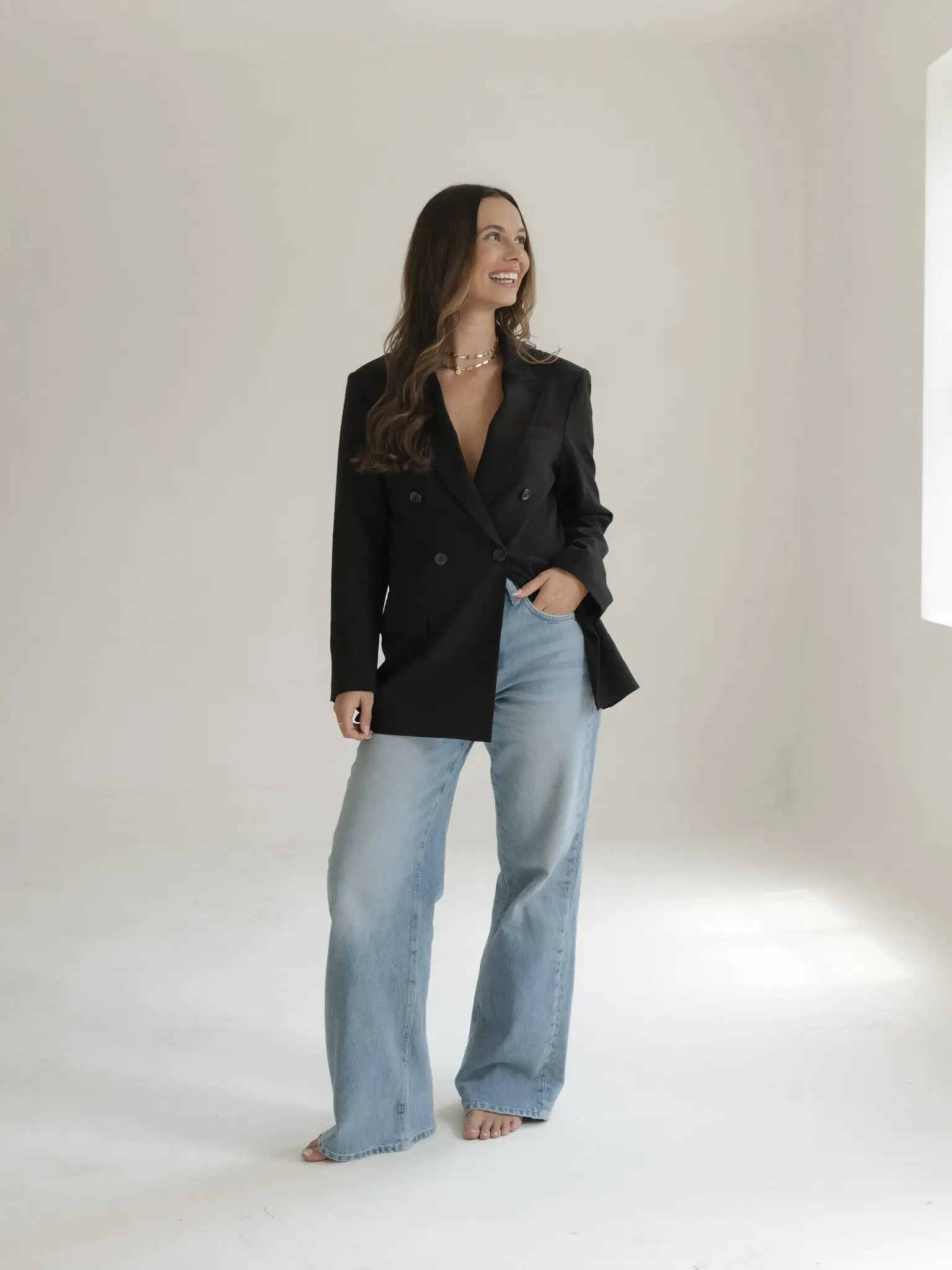 Paige Pope standing in a bright studio in a black blazer and jeans, smiling toward the window