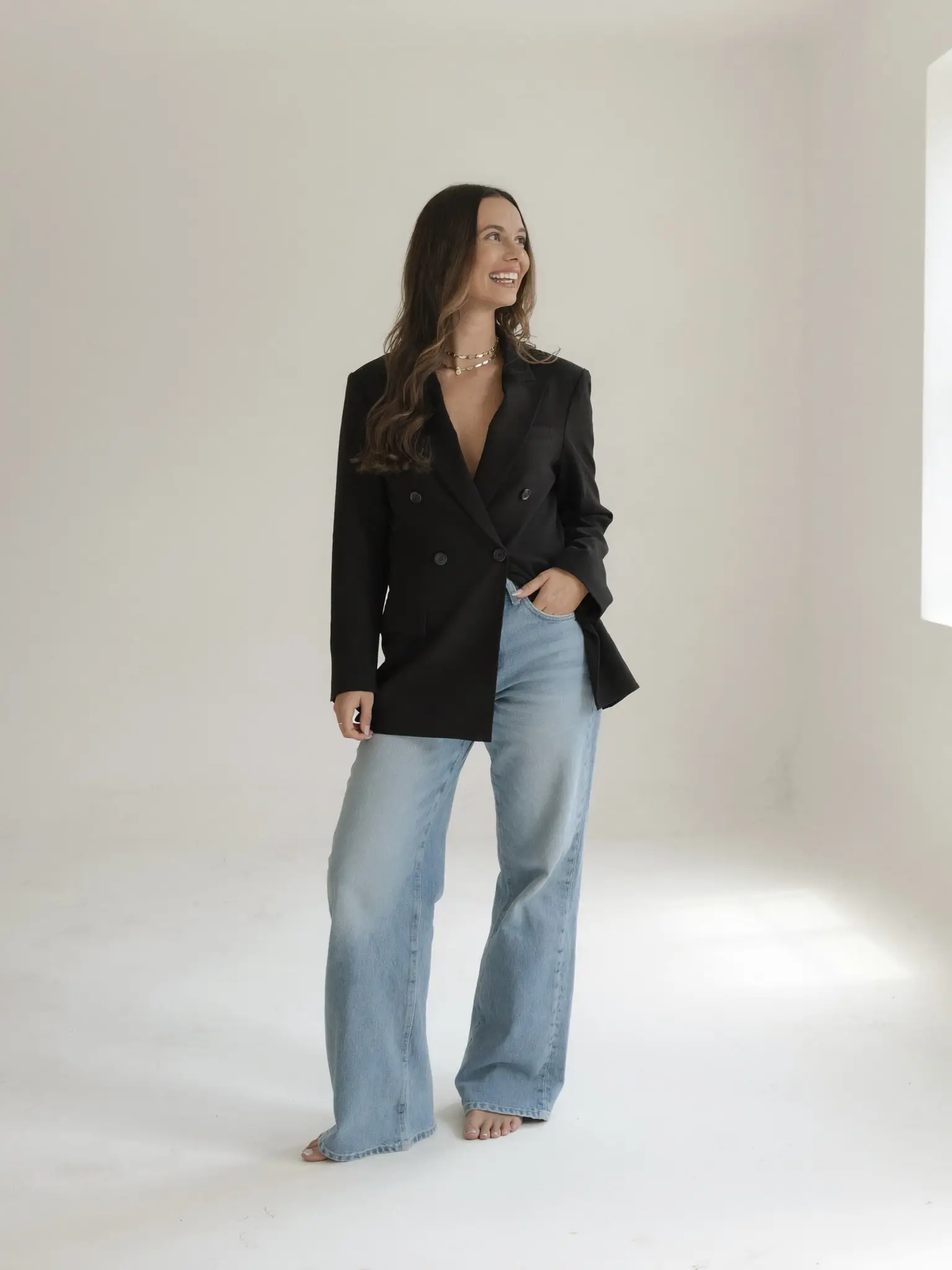 Paige Pope standing in a bright studio in a black blazer and jeans, smiling toward the window