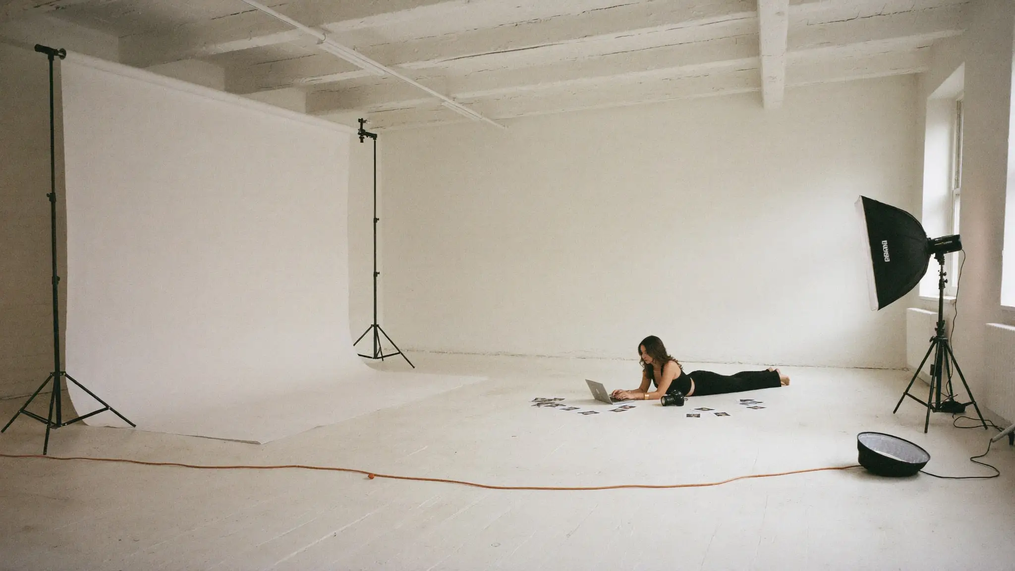 Paige Pope working on a laptop on the floor of a large white photo studio, between a paper backdrop and a softbox light